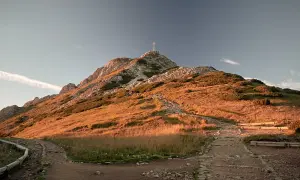 Giewont z krzyżem na szczycie, oświetlony zachodzącym słońcem. Ścieżka prowadzi w górę, a w oddali widać ławki dla turystów.