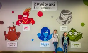 Dwie dziewczynki pozują na tle ściany z postaciami z bajki "Żywiołaki Elementoons". Widać Ogi Blaze, Ziemka Rocky, Wodzię Flow, Aero Airy i Żywka Zippy.