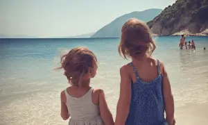 Dwie dziewczynki trzymają się za ręce na plaży, patrząc na morze. W tle widać góry i innych ludzi kąpiących się w wodzie. To idealne wakacje w Albanii z dziećmi.