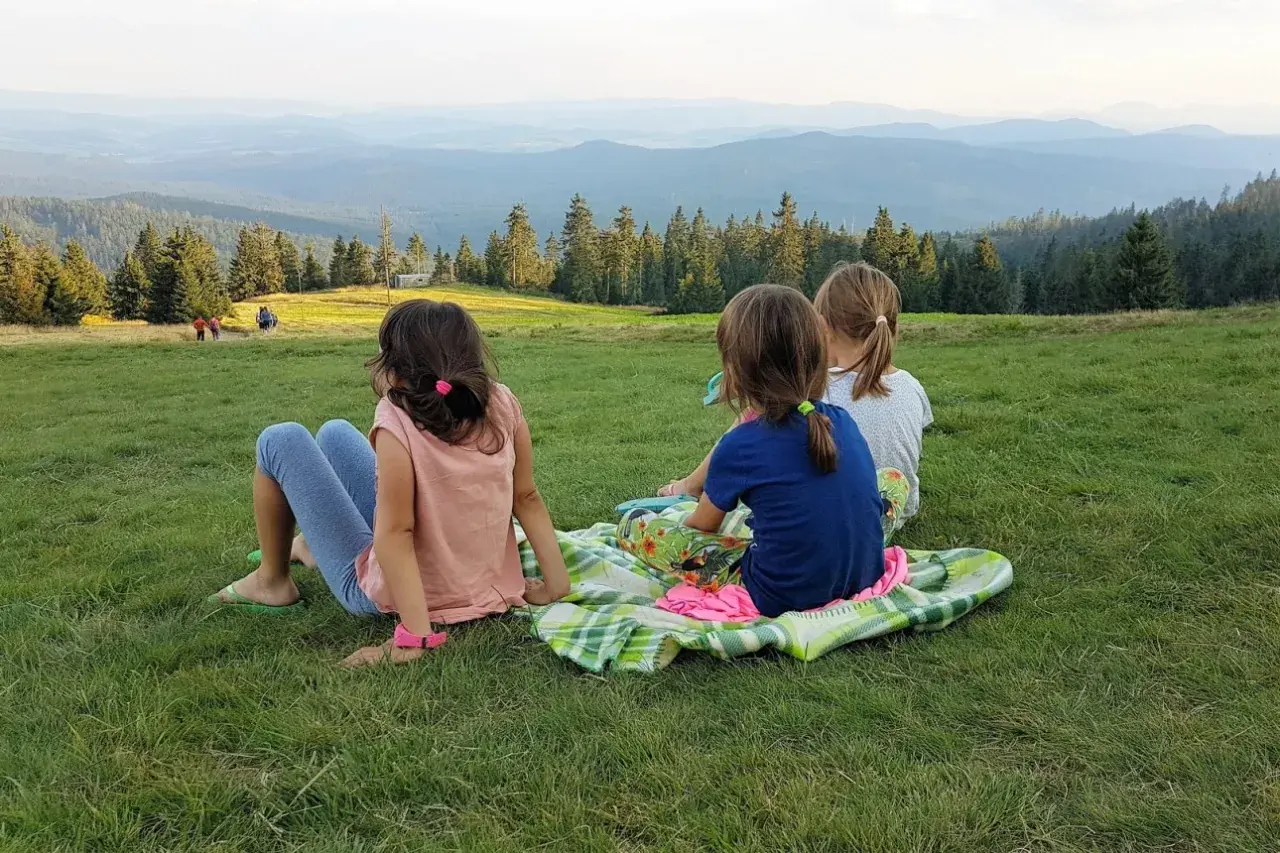 Trzy dziewczynki siedzą na kocu na trawie, podziwiając rozległy widok na Beskid Żywiecki.
