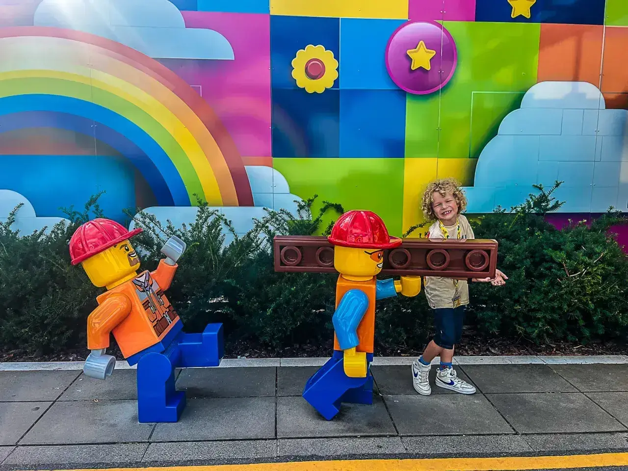 Chłopiec z uśmiechem pozuje z dwoma wielkimi ludzikami LEGO na tle kolorowego muralu. To świetne billund atrakcje dla całej rodziny!