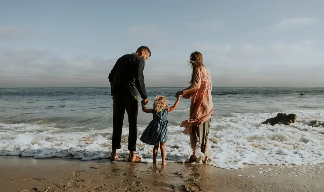 Rodzina na plaży, szczęśliwe wakacje z niemowlakiem. Dziecko trzyma rodziców za ręce, fale obmywają ich stopy.