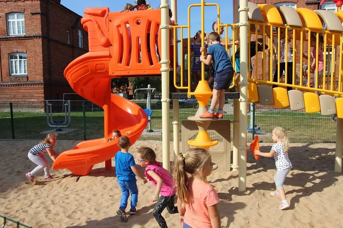 Kolorowy plac zabaw z pomarańczową zjeżdżalnią i żółtymi elementami, gdzie dzieci w różnym wieku, w tym maluchy, cieszą się zabawą. Idealne atrakcje dla 3 latka.
