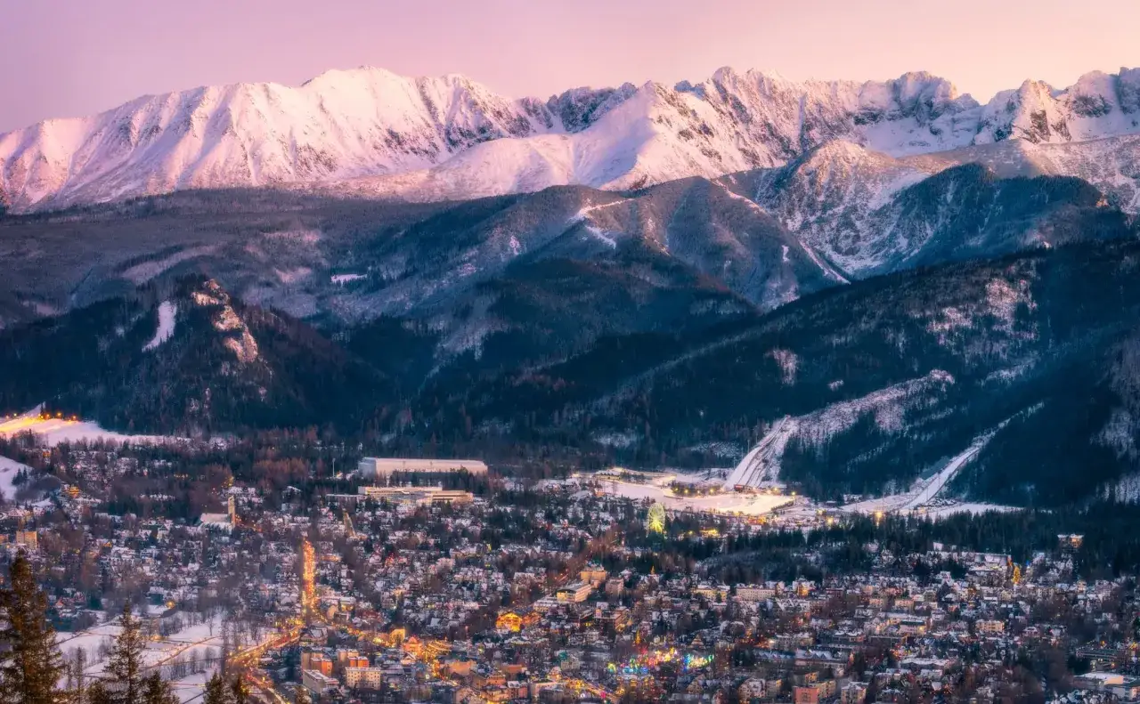 Ośnieżone Tatry nad rozświetlonymi Zakopanem. Widoczne skocznie narciarskie i koło widokowe, zapowiadające zimowe atrakcje.