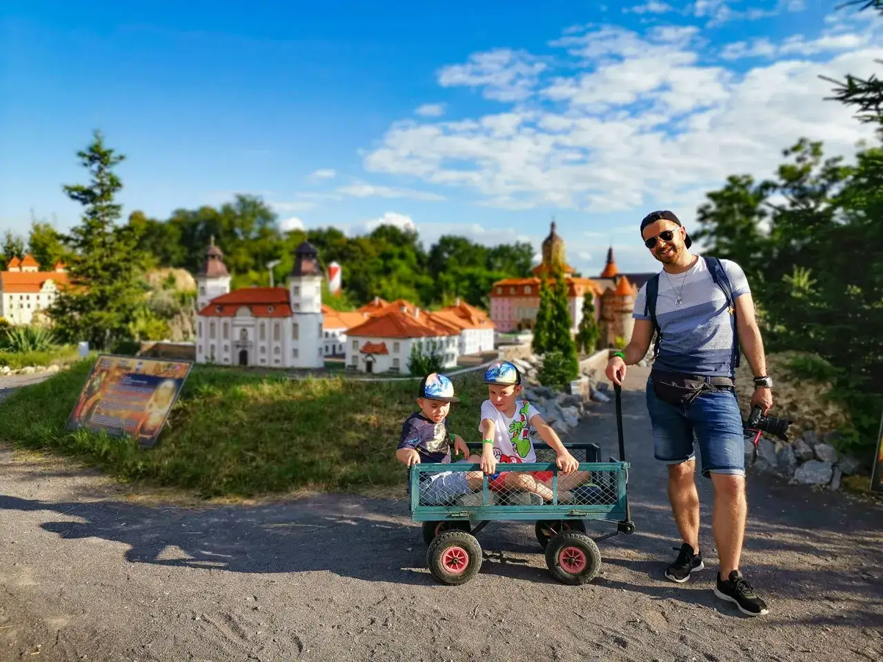 Ojciec z synami zwiedza mini city. Dzieci siedzą w wózku, a tata z aparatem w ręku uśmiecha się do obiektywu.