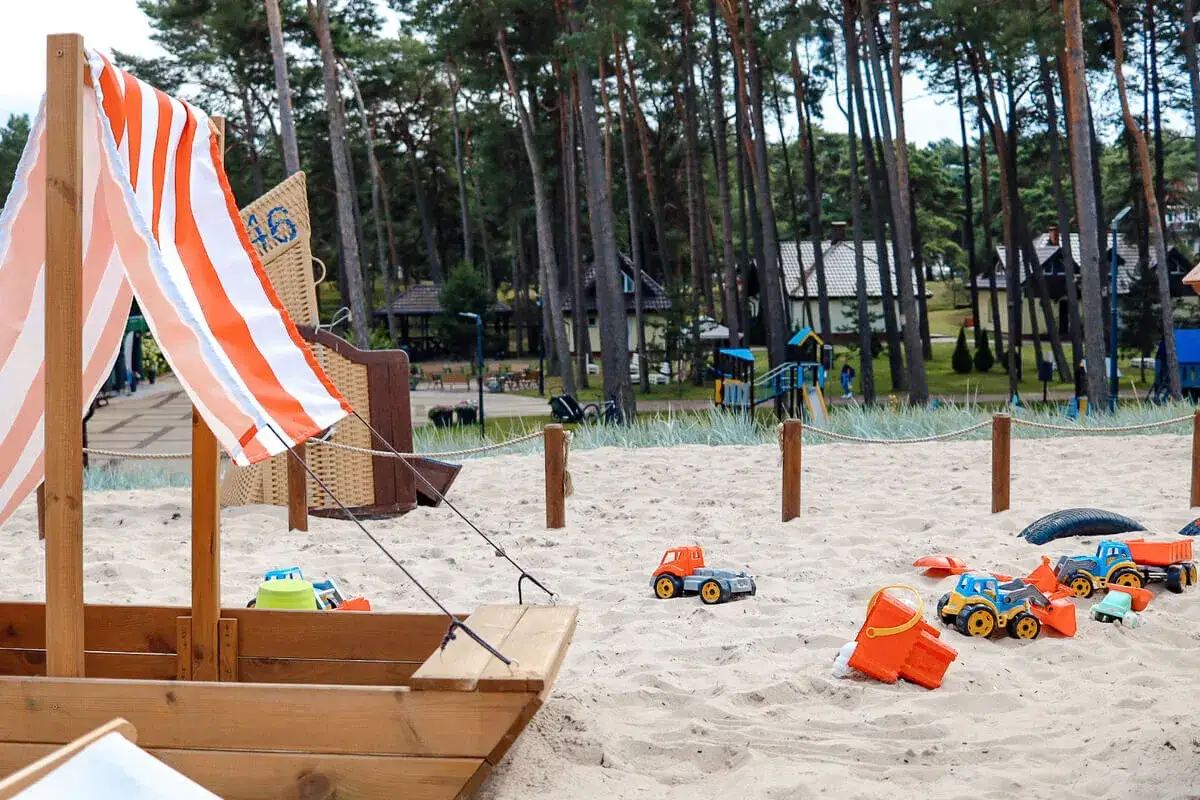 Piaskownica z zabawkami i łódką zachęca do zabawy. W tle plac zabaw i las, idealne ustka atrakcje dla dzieci.