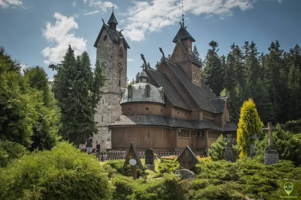 Drewniany kościółek i kamienna wieża z zegarem, otoczone zielenią i nagrobkami. Jedna z głównych karpacz atrakcji.