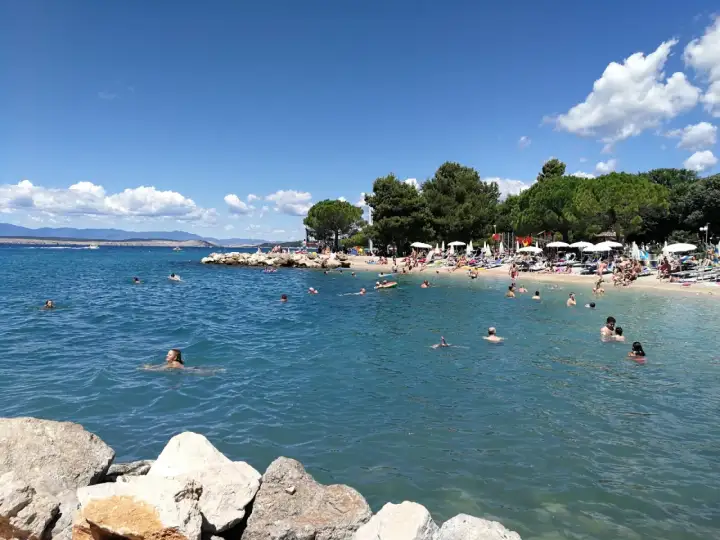 Crikvenica atrakcje: plaża pełna ludzi kąpiących się i opalających, błękitne morze, zielone drzewa i góry w tle.